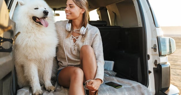 Large white dog and woman sitting in the back of a car on a blanket with the door open.