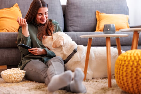 Smiling woman sitting on a living room floor with a bowl of popcorn while a Labrador lies beside her.