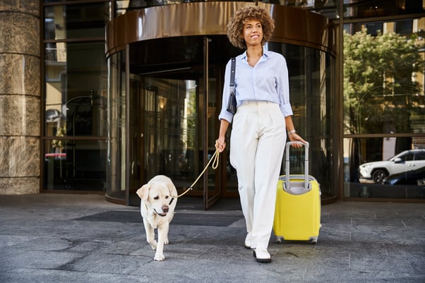 Woman leaving a hotel rolling a bright yellow suitcase, with her dog on a lead alongside her.