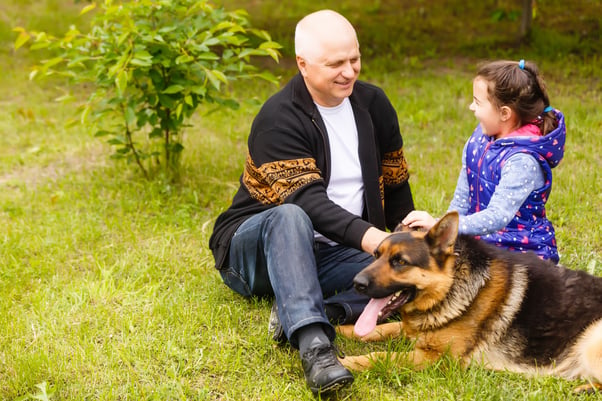 Dad and daughter sitting on the grass gently petting a German Shepherd dog.