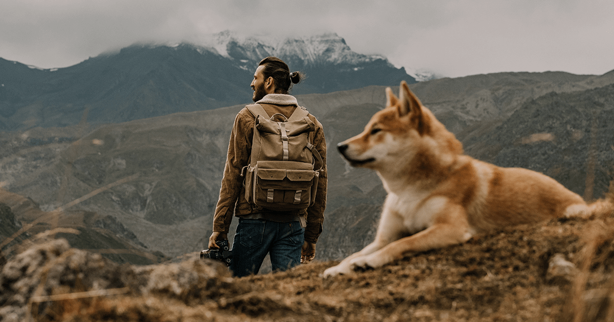 Man looking off at mountains in the distance with a camera in hand while Shiba Inu dog lies in the foreground