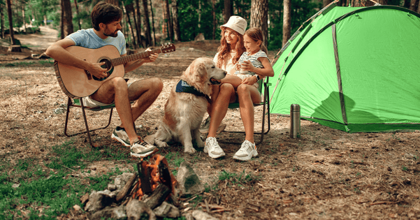 Young family sitting around campfire with dog while dad plays guitar