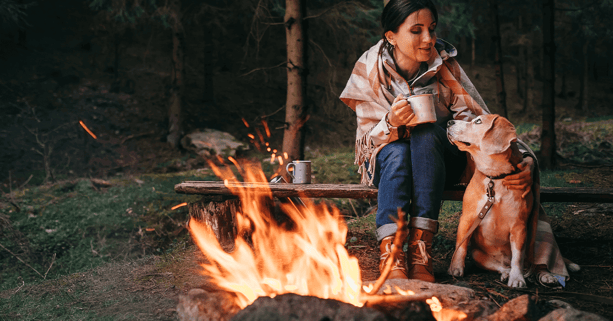 Woman sitting by fire with a mug and blanket and petting her dog