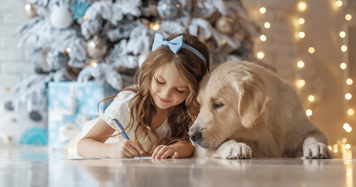 Little girl laying in front of Christmas tree writing letter to Santa beside Golden Retriever