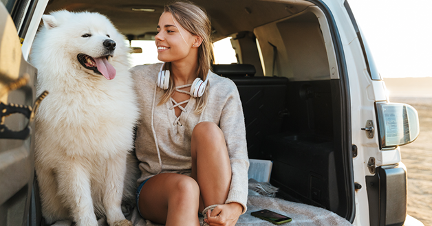 um  grande cão branco e uma mulher sentados sobre um cobertor no porta-malas de um carro, com a porta aberta